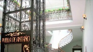 Une porte en fer forgé richement décorée, sur laquelle on peut lire l’inscription « Plaza de Antino ». À l’intérieur, un escalier à vis s’élève, entouré de meubles en bois, et offre une vue sur l’espace intérieur.