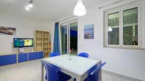 A modern dining area with a glass table and blue chairs, a television on a cabinet, and a large window with a view of the outdoors. - Scirocco, Holiday Home with garden and terrace ,100 mt from the sandy beach (Marina di Ragusa)