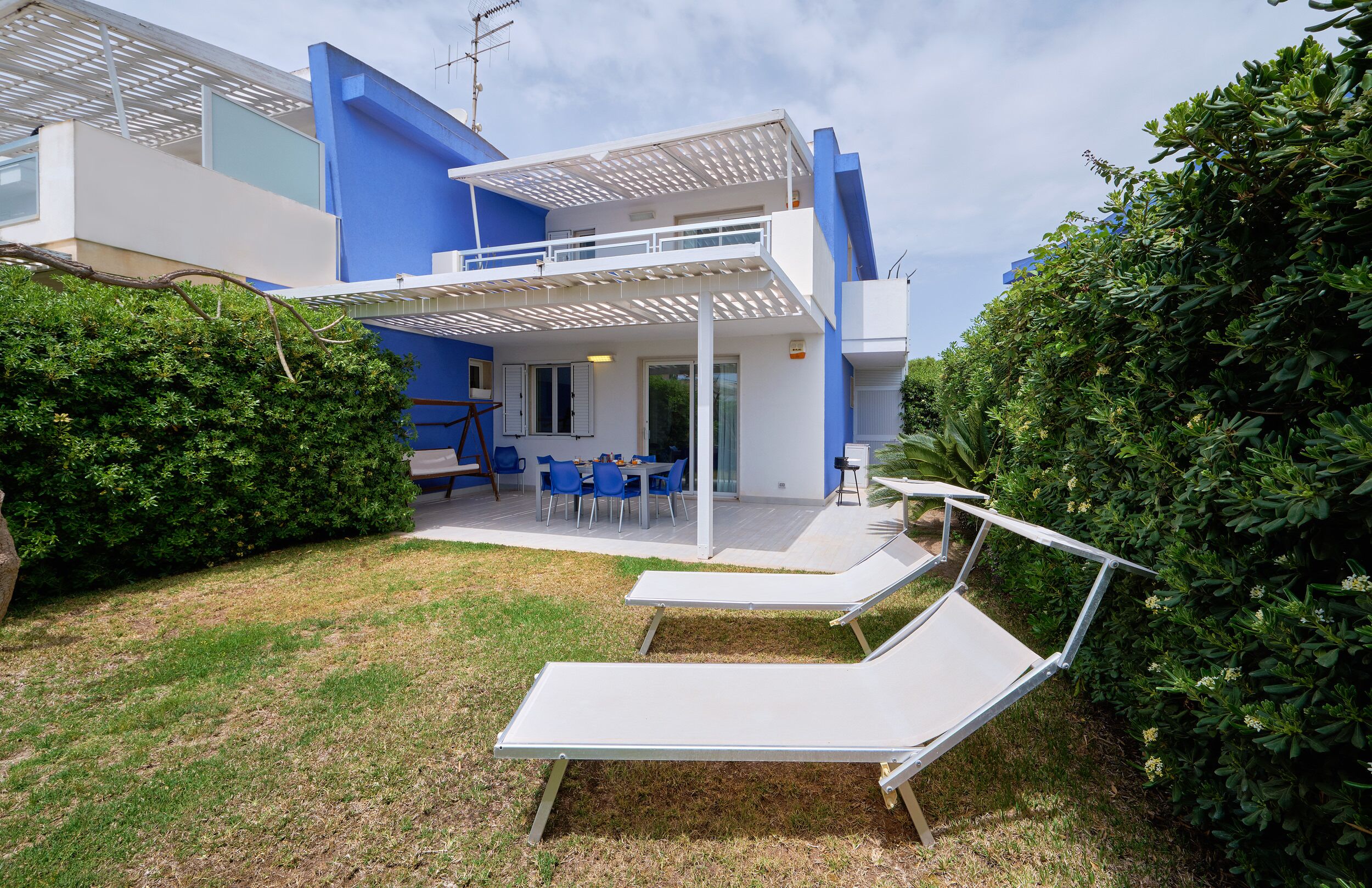 A modern two-story house with a blue exterior, a covered patio with outdoor furniture, and a well-maintained lawn.