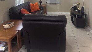 Un salon avec un canapé en cuir noir, un pouf, une table basse en bois, un téléviseur fixé au mur et un ventilateur de plafond.