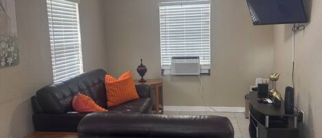 A living room with a black leather sofa, an ottoman, a wooden coffee table, a TV mounted on the wall, and a ceiling fan.
