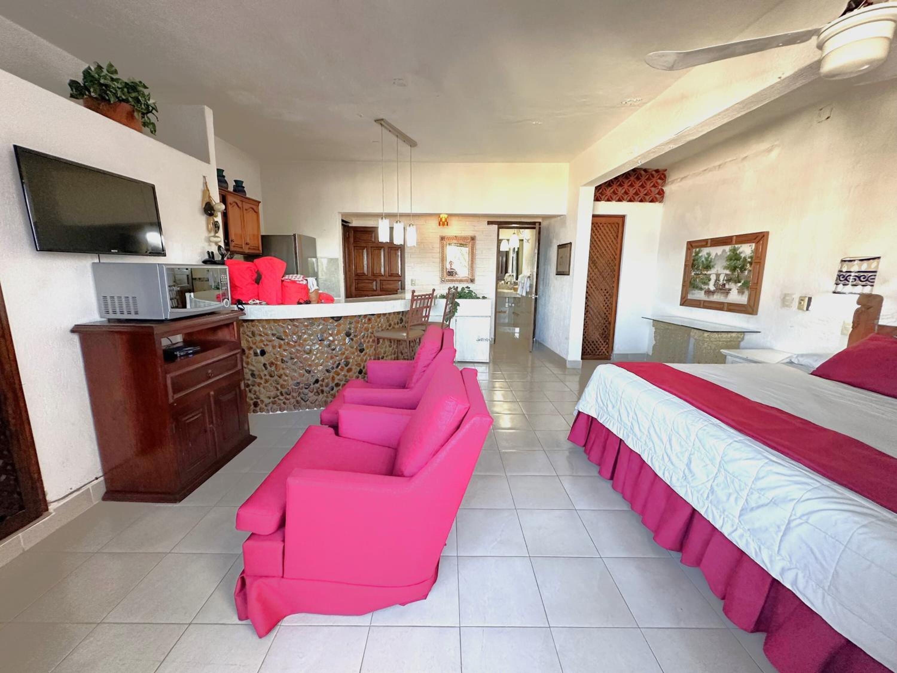 A hotel room with a kitchenette, a TV, a pink sofa, a bed with a red bedspread, and a ceiling fan.