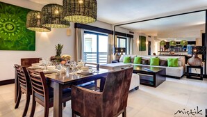 Ein modernes Wohnzimmer mit Essbereich, ausgestattet mit Korbtreleuchten, einem großen, grünen abstrakten Gemälde und Blick auf die Küche und den Balkon.
