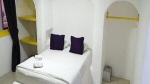 Una cama individual con colchón blanco y dos almohadas oscuras, una mesita de noche con una toalla blanca y un perchero amarillo.