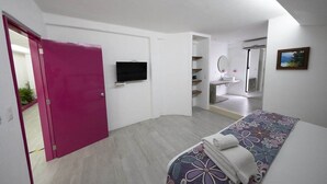 Un dormitorio moderno con una puerta rosa, un televisor de pantalla plana, una cama con colcha estampada y un baño con espejo y lavabo.