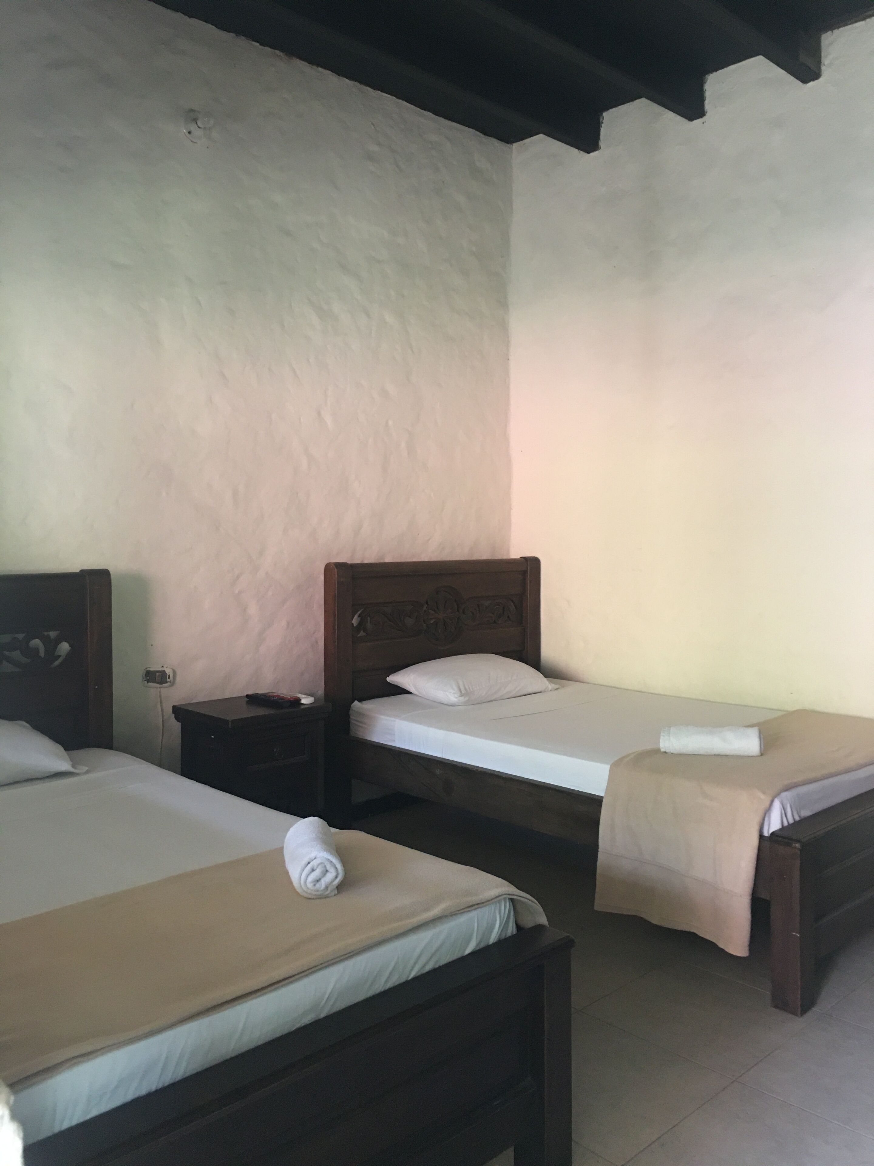 Una habitación con dos camas de madera, cada una con un colchón blanco y una manta color beige. Entre las camas hay una mesita de noche con una lámpara.