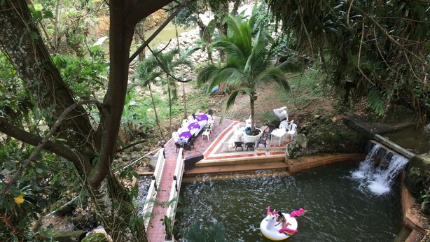 Uma piscina natural com cascata, rodeada por vegetação exuberante e uma área de estar com cadeiras e mesas.