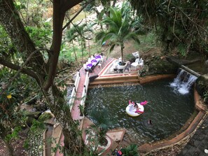 Ein natürlich angelegter Pool mit Wasserfall, umgeben von üppiger Vegetation und einer Sitzgelegenheit mit Stühlen und Tischen.