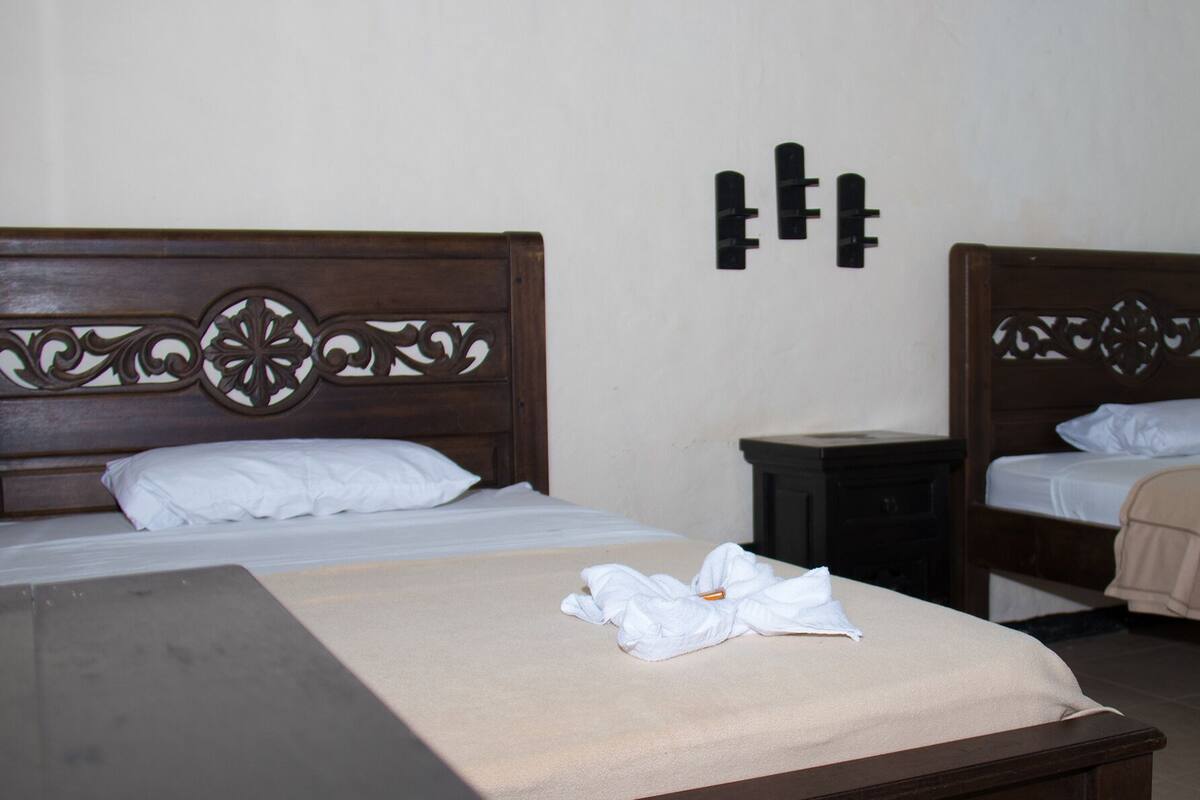 Ein Hotelzimmer mit zwei hölzernen Betten, einem kleinen Nachttisch und einem großen Bett mit einer beigefarbenen Decke.