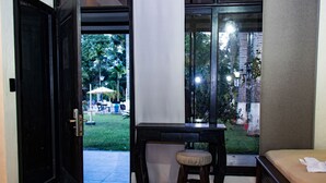 Quarto com cama, escrivaninha e vista para área externa com gramado e guarda-sóis.