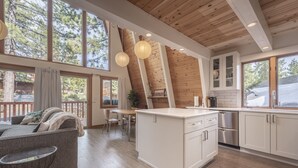 Una cocina moderna con vigas de madera, un área de comedor con sofá y mesa, y grandes ventanales con vistas a árboles.