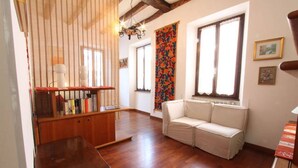 A room with wooden flooring, a white sofa, a wooden table, a bookshelf, and a window with a floral curtain. - Captivating 4 -bed Apartment in Bellagio Historic (Bellagio)