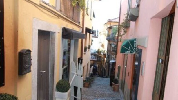 A narrow alleyway with buildings on either side, a door on the left, and a small white chair in the center. - Captivating 4 -bed Apartment in Bellagio Historic (Bellagio)