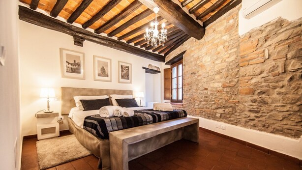 Un dormitorio con pared de piedra, techo de madera, cama con almohadas, un banco, una mesita de noche y una lámpara.