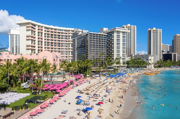 A beachfront hotel with multiple high-rise buildings, a sandy beach with umbrellas and lounge chairs, and palm trees.