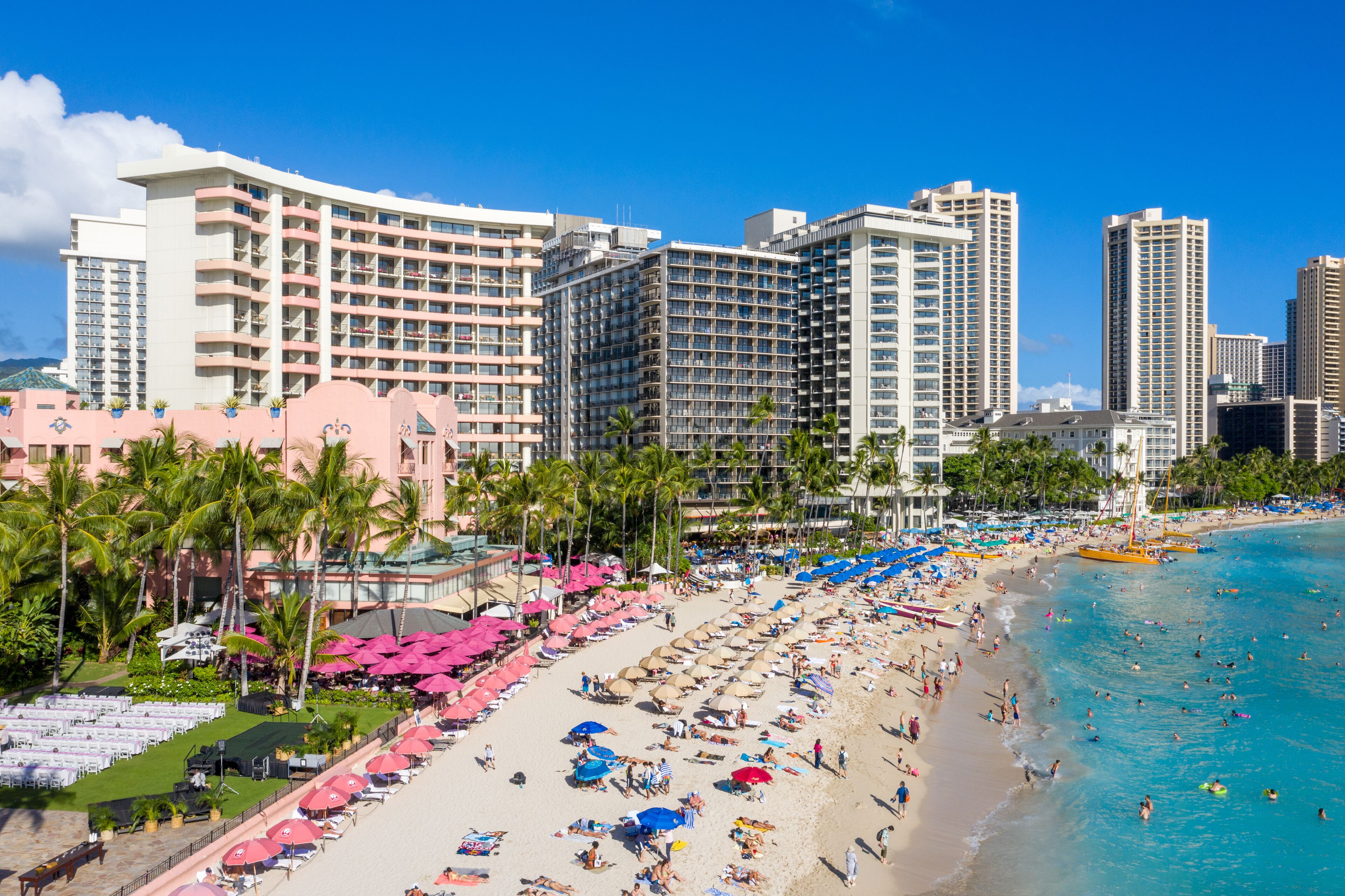 A beachfront hotel with multiple high-rise buildings, a sandy beach with umbrellas and lounge chairs, and palm trees.