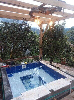 Een hottub onder een houten pergola, omringd door weelderige begroeiing en een bergachtige achtergrond.