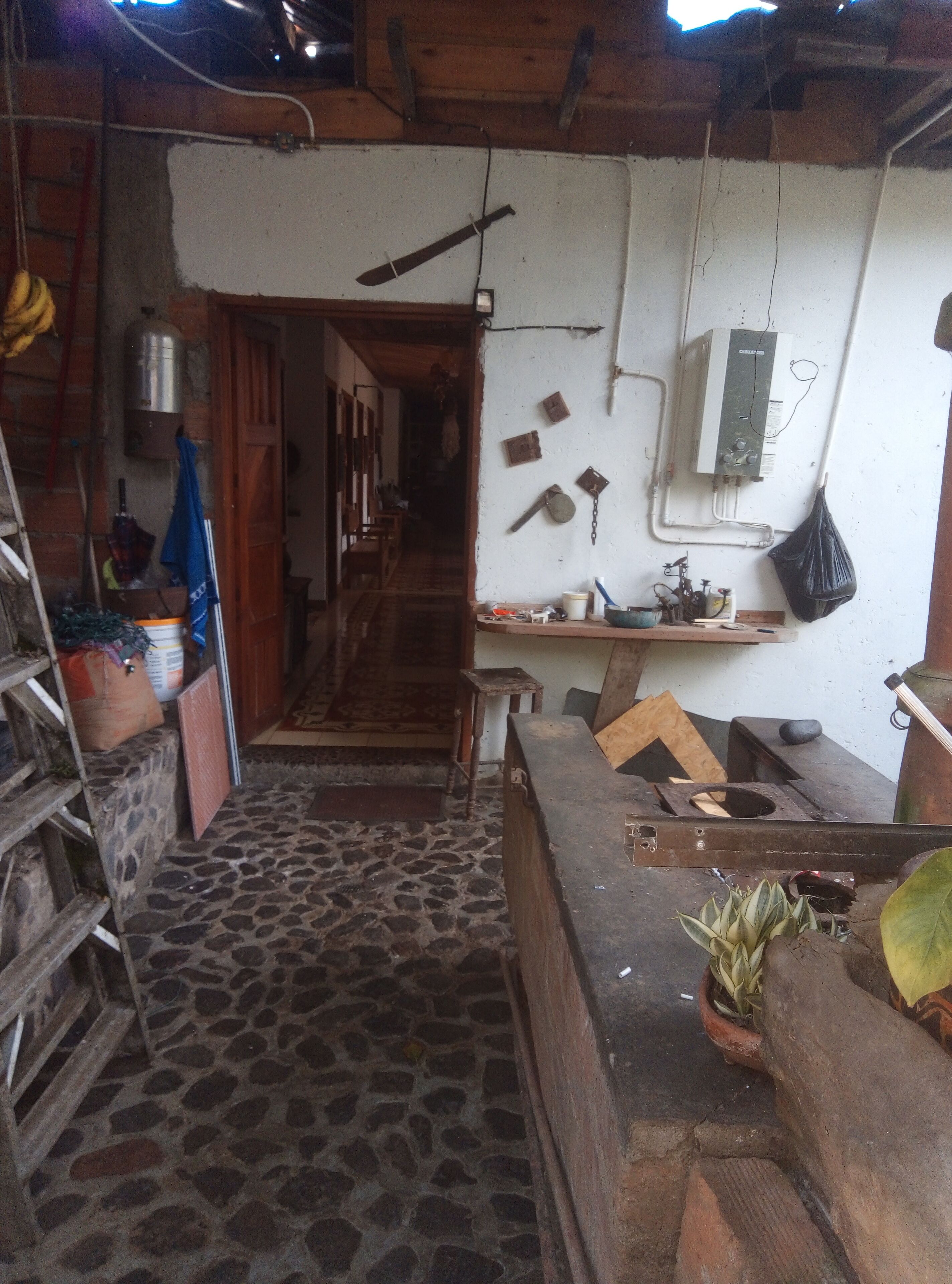 A rustic kitchen with a stone floor, wooden beams, and basic amenities including a sink, stove, and storage shelves.
