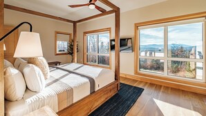 A bedroom with a large bed, wooden beams, a ceiling fan, and multiple windows offering a view of the outdoors.