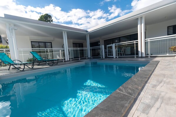 A swimming pool with a tiled edge, surrounded by a covered patio area with white railings and a few lounge chairs.