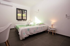 A bedroom with a bed, a chair, a bedside table with a lamp, a window with blinds, and an air conditioner on the wall.