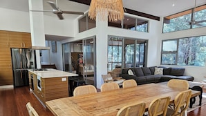 A spacious kitchen with a wooden dining table and chairs, a refrigerator, and a large window overlooking a forested area.