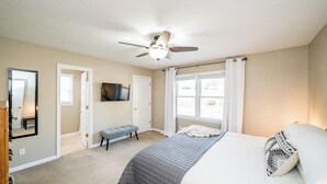 Ein Schlafzimmer mit einem Bett, einem Deckenventilator, einem Fernseher und einem Fenster mit Vorhängen.