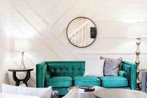 Een smaragdgroene bank met twee kussens, een ronde spiegel en twee lampen aan weerszijden.