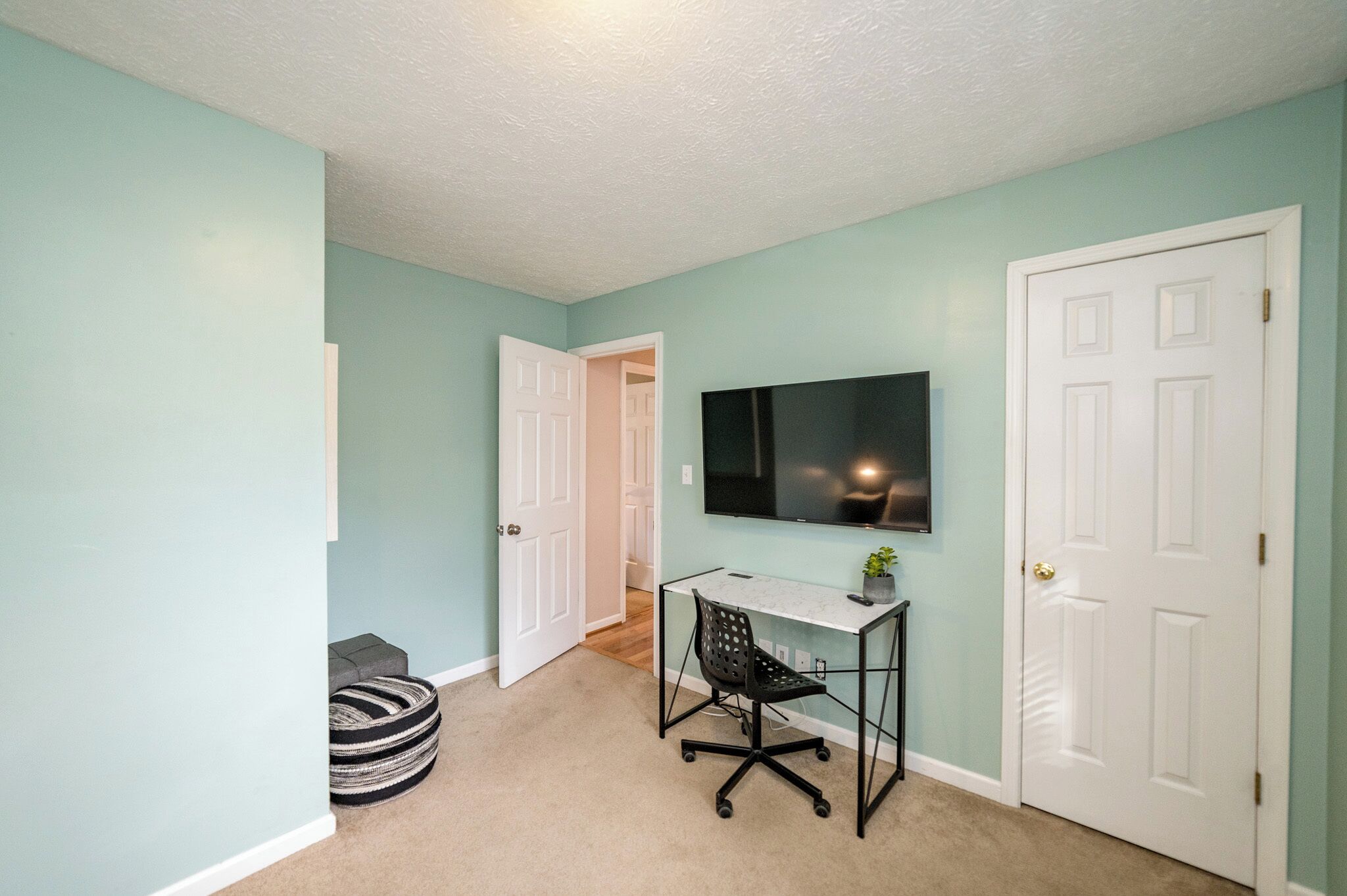 A hallway with teal walls, a white door, a black office chair, a small table, and a TV mounted on the wall.