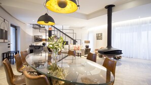 Een moderne woonkamer met een eettafel met glazen blad, omringd door lederen fauteuils, een zwarte open haard en eigentijdse decoratie.