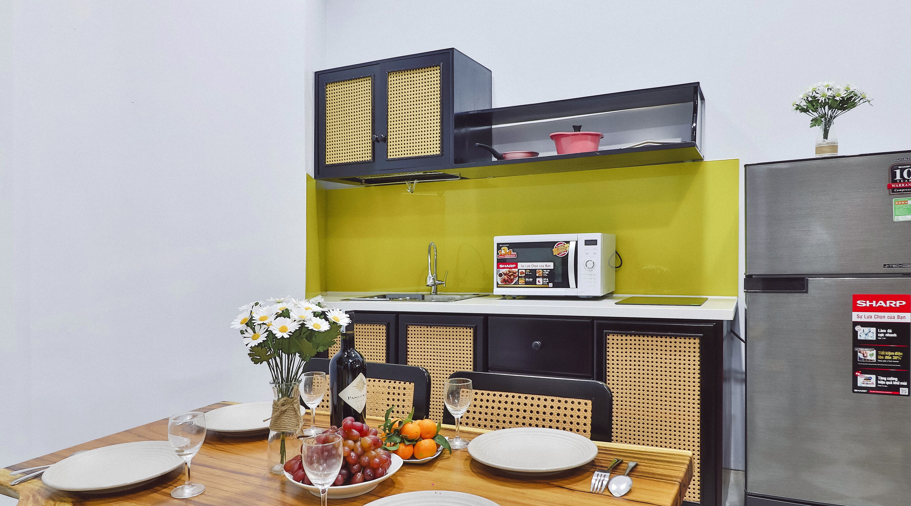 Ett kompakt kök med mikrovågsugn, ett kylskåp från Sharp och ett matbord med tillhörande tallrikar, glas och en fruktskål.