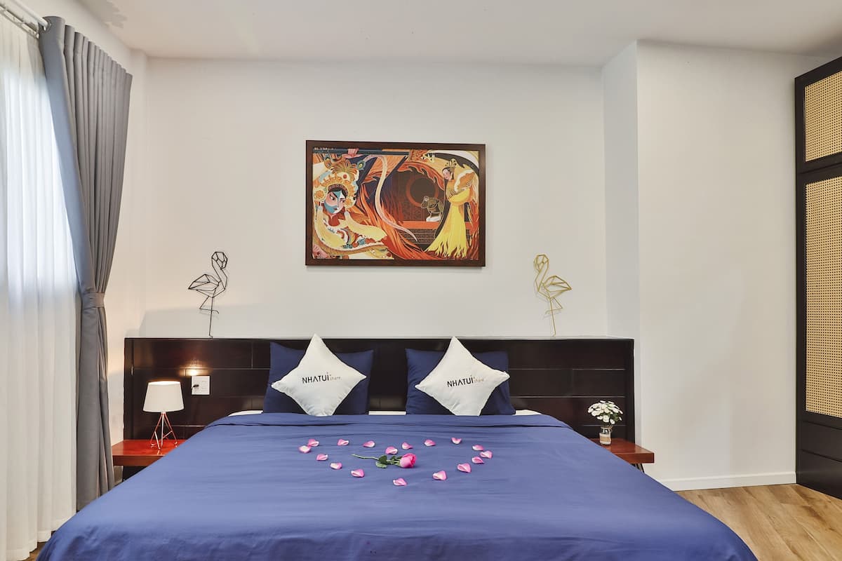 A bedroom with a bed, a nightstand, a lamp, a painting, and a window with curtains.