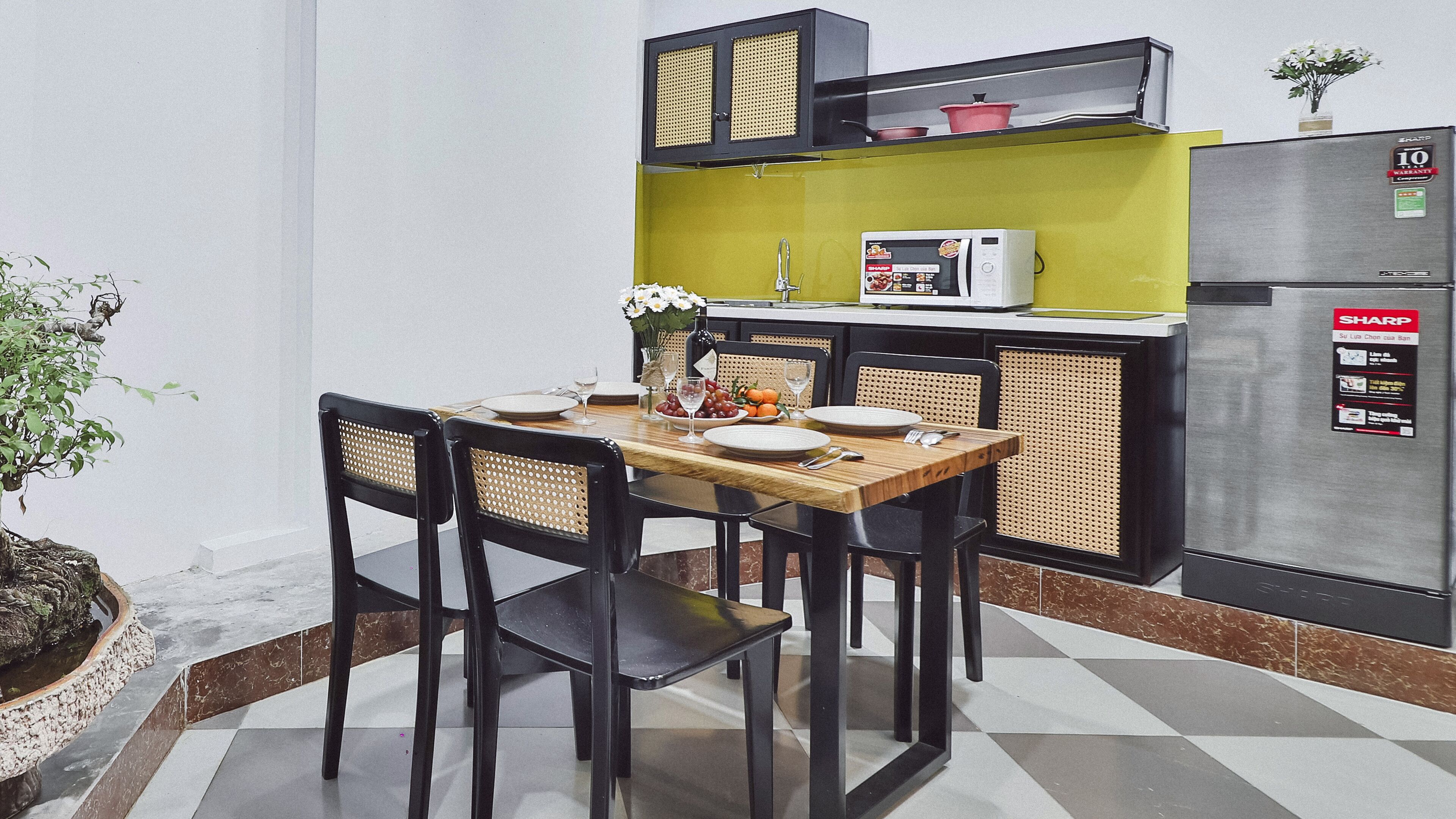 Een kleine keuken met een eettafel voor vier personen, een magnetron, een koelkast en een klein plankje met keukengerei.