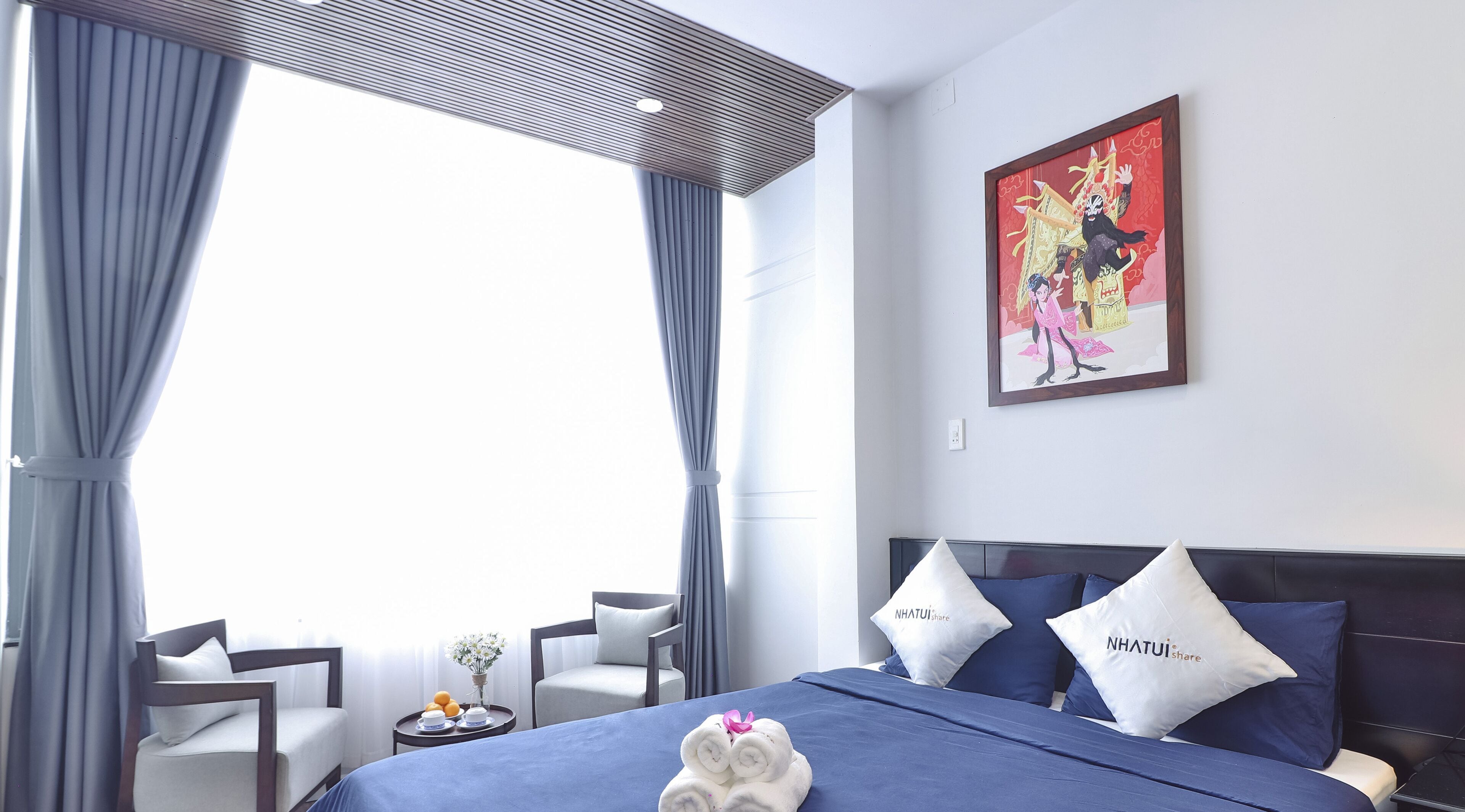 A hotel room with a large bed, two chairs, a small table with a vase and oranges, and a painting on the wall.