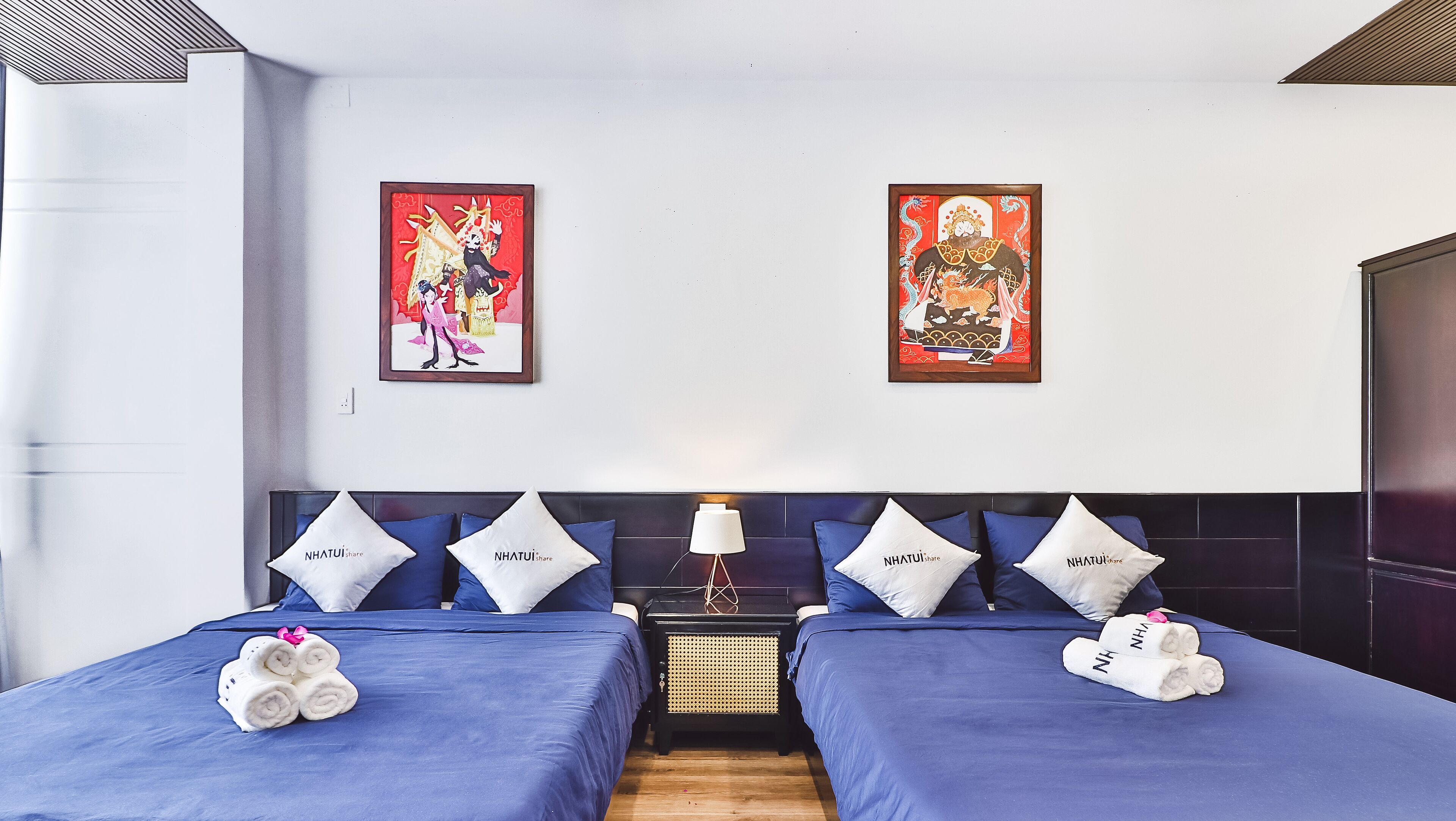 Une chambre d’hôtel avec deux lits, chacun agrémenté de draps et d’oreillers bleus portant l’inscription « Nhatui share ». Deux œuvres d’art encadrées ornent le mur, ainsi qu’une petite table avec une lampe et des serviettes sur les lits.