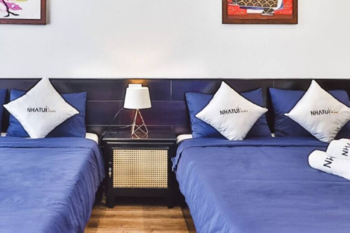 A hotel room with two beds, each with blue bedding and pillows labeled "NHATUI." There are towels on the beds and a small table with a lamp between the beds.