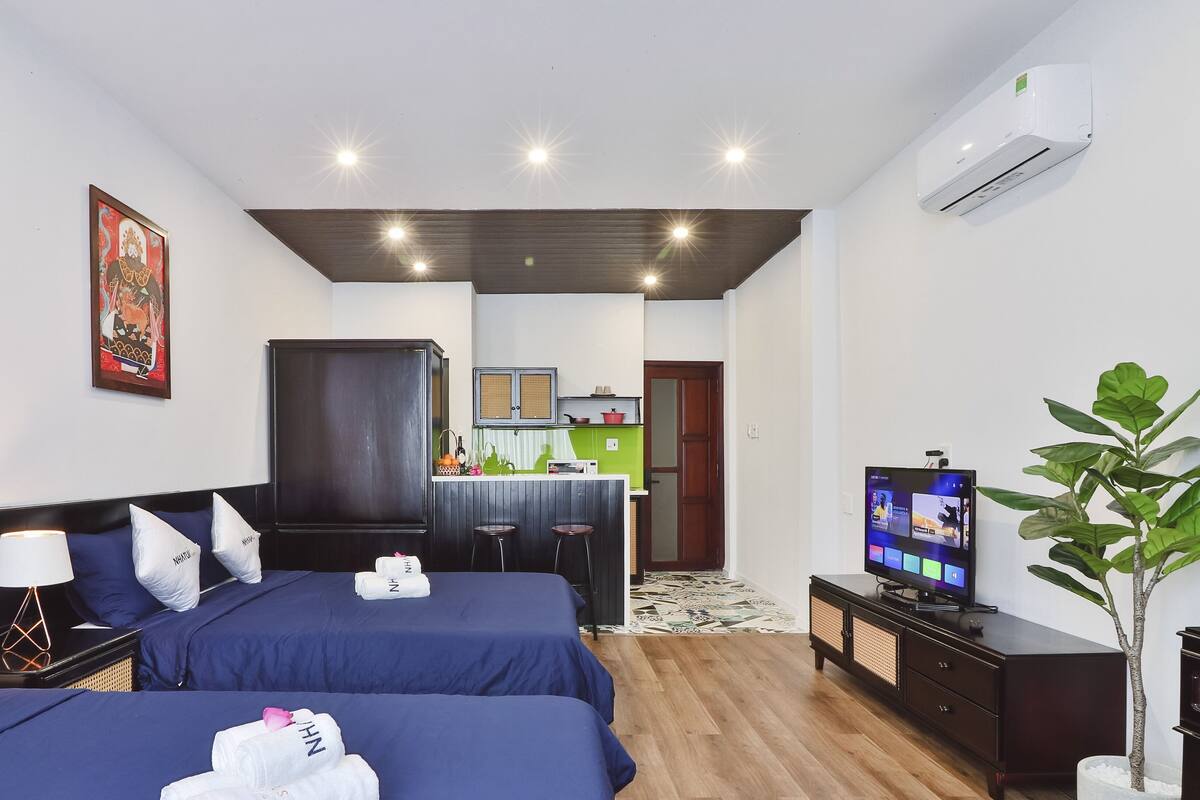 A hotel room with two beds, a TV, a desk, a chair, a plant, and a kitchen area in the background.