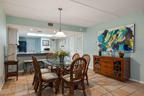 Uma área de refeições com uma mesa de vidro rodeada por cadeiras de vime, um armário de madeira com gavetas e um grande quadro abstrato na parede.