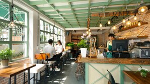 Un café au décor rustique, avec un mur en briques apparentes, des poutres vertes au plafond et des ampoules suspendues. On y trouve des tables et des chaises en bois, et une personne se tient au comptoir.