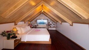 Une chambre avec un plafond en pente, un lit, une table de chevet avec une lampe, et une vue sur le paysage urbain par la fenêtre.