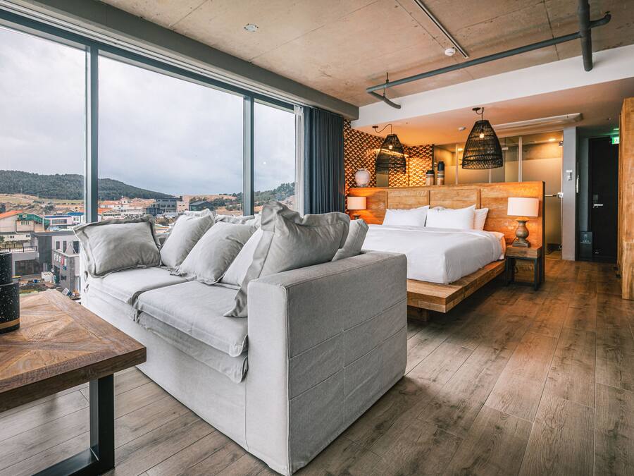 A modern hotel room with a large window, a sofa, a bed, and a wooden table.