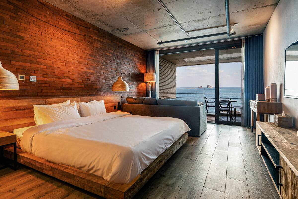 A modern hotel room with a large bed, a sofa, a dining area, and a view of the sea.