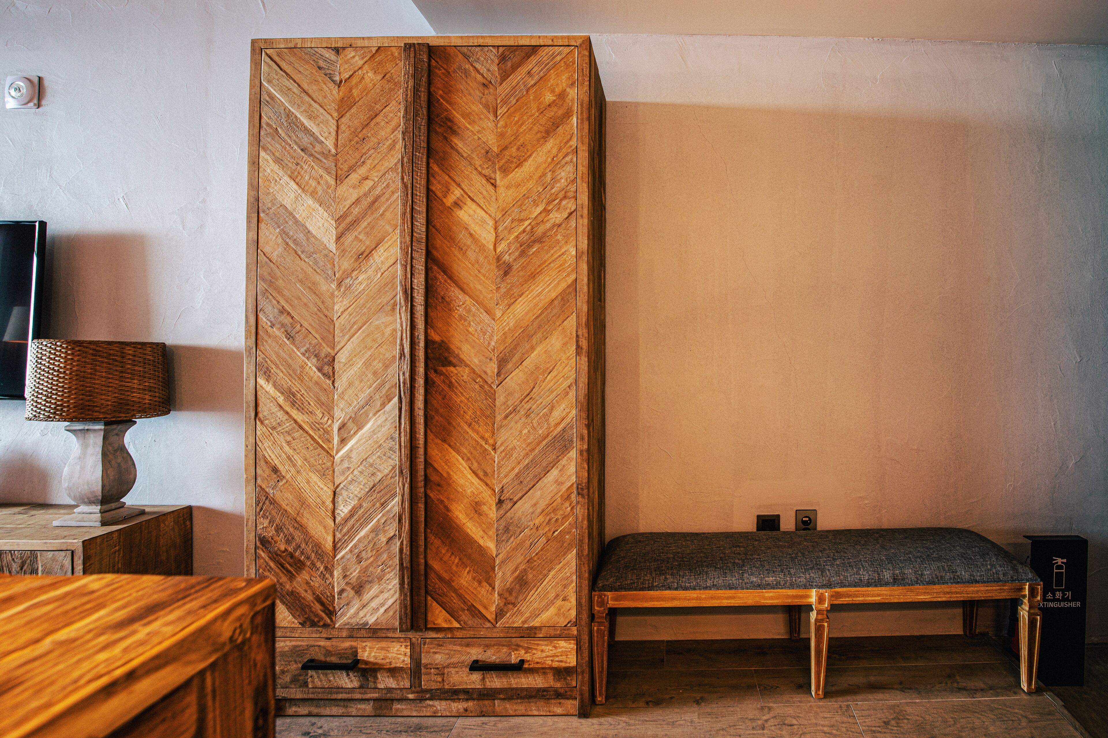 A room with a herringbone-patterned wooden wardrobe, a bench with a cushion, and a lamp on a side table.