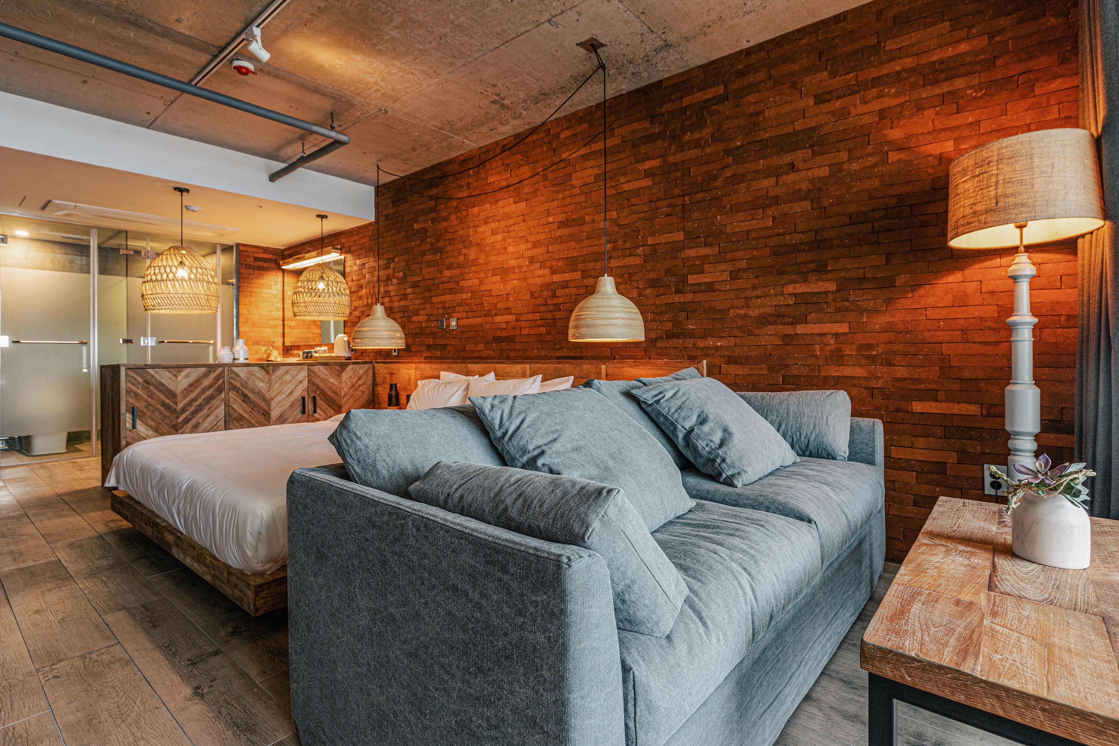 A modern hotel room with a brick wall, a grey sofa, a wooden coffee table, and pendant lighting.