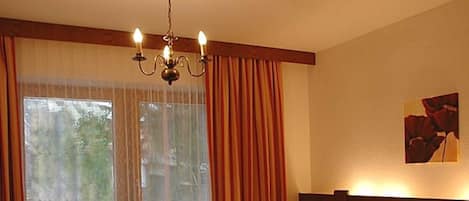 A hotel room with a bed, a chandelier, a window with curtains, and a nightstand with a vase of flowers.