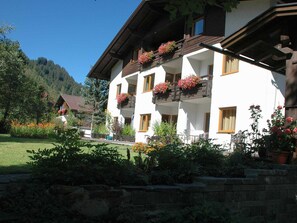 A white building with balconies, surrounded by greenery and a mountain backdrop.