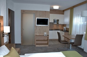 Quarto de hotel com cama, mesa de cabeceira, luminária, TV, cozinha compacta e área de refeições com cadeiras.
