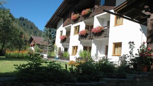 A white building with balconies, surrounded by greenery and a mountain backdrop. - TAN 1 east (1-2 persons) - Apartments Vilsalp (Bogen)