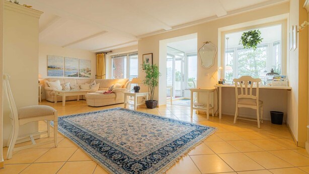 Uma sala de estar ampla com um tapete azul estampado, um sofá, uma mesa de jantar com cadeiras e vista para o exterior por grandes janelas.