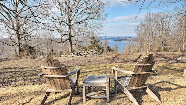 Zwei Adirondack-Stühle und ein kleiner Holztisch stehen auf einer Wiese mit Blick auf einen See und Bäume.